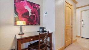 A room with a wooden desk, a chair, a lamp, a painting of flowers, and a door. - Lodge Tower 782 Lodge Room (Vail)