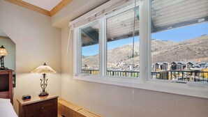 A bedroom with a large window, a bedside lamp, a wooden nightstand, and a view of a mountain and buildings. - Lodge Tower 782 Lodge Room (Vail)