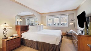 A hotel room with a large bed, a wooden dresser, a desk, and a television. There are windows with a view of a mountainous landscape. - Lodge Tower 782 Lodge Room (Vail)