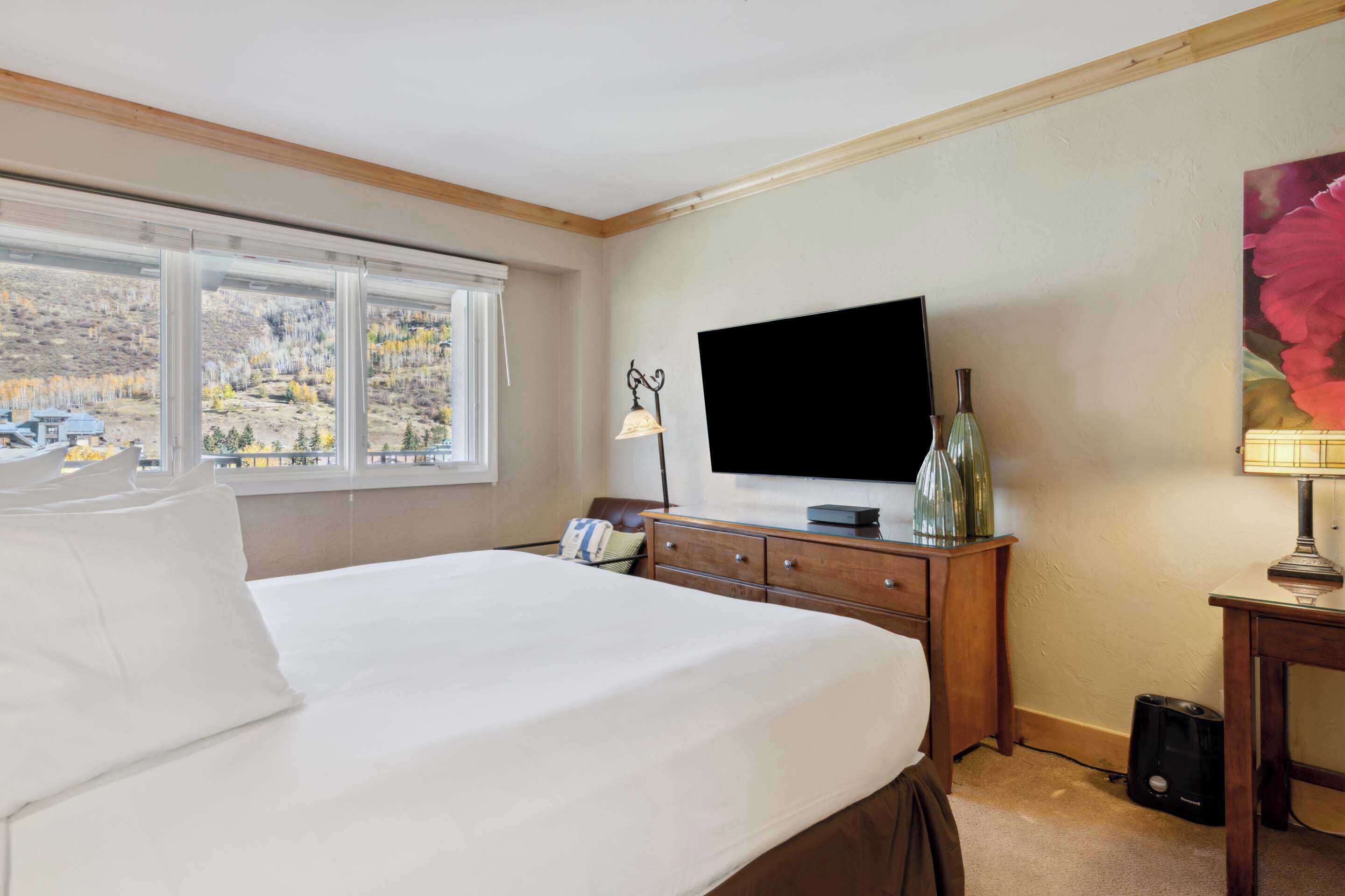 A hotel room with a large bed, a television, a wooden dresser, and a window with a view of a mountainous landscape.