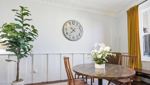 A dining area with a round wooden table, chairs, a potted plant, a wall clock, and a window with curtains. - Cozy 2BR Loft in Downtown Charleston, IL (Charleston)