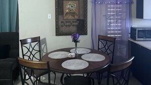Un comedor con una mesa redonda, cuatro sillas, un jarrón con flores, un cuadro en la pared, una ventana con cortinas y un microondas en la encimera.