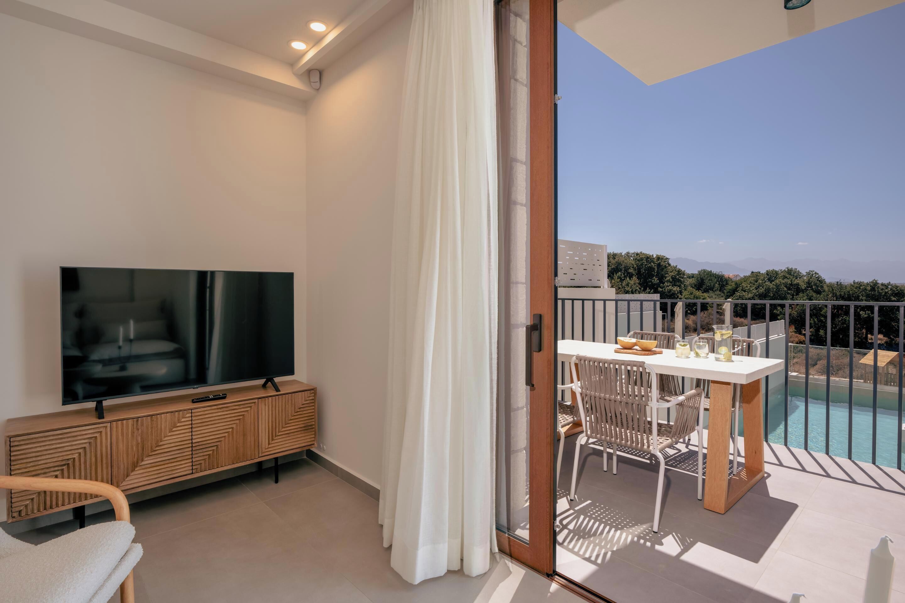A modern living room with a flat-screen TV, wooden cabinet, and a balcony with a table and chairs overlooking a pool and mountains.
