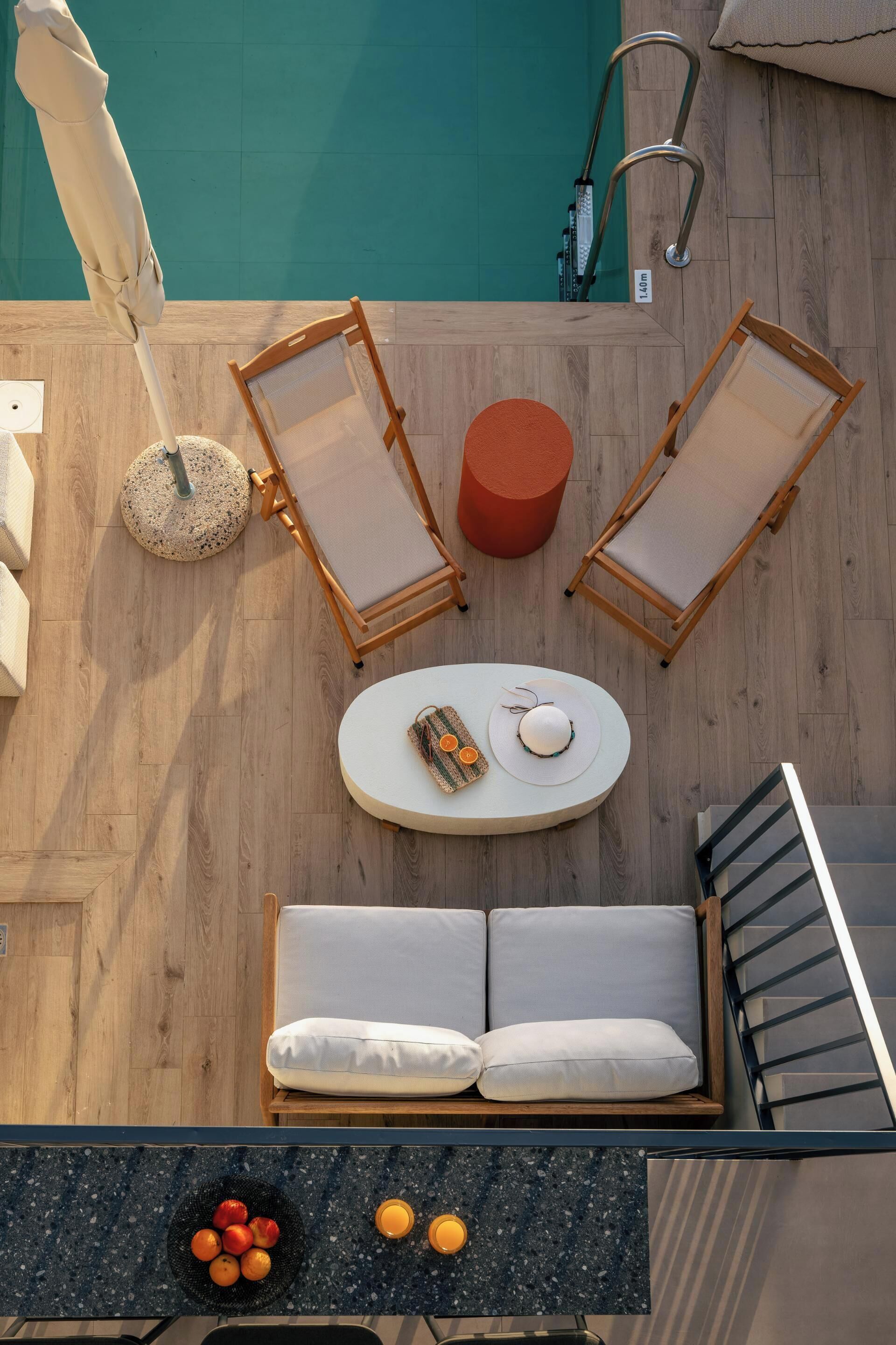 A poolside lounge area with two lounge chairs, a round table with a hat and fruit, a small orange stool, and a sofa with cushions.