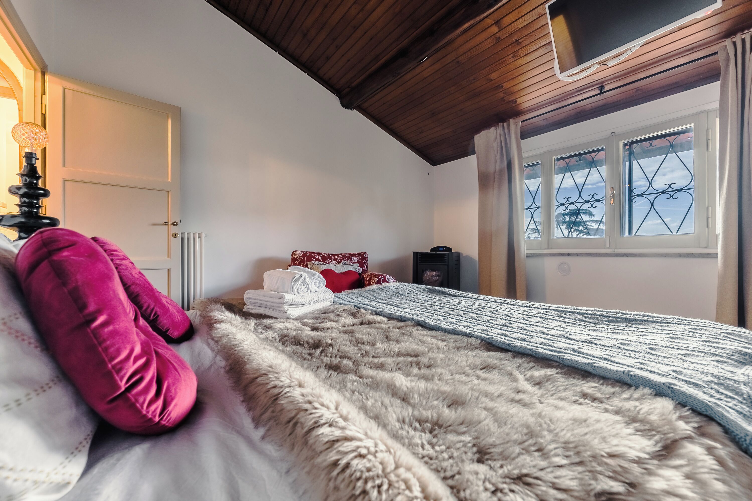 A bedroom with a wooden ceiling, a bed with a fur throw, a window with metal bars, and a radiator.