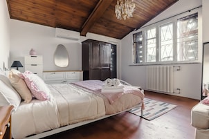 Un dormitorio con techo de madera, cama con ropa de cama rosa y blanca, cómoda, espejo y ventana con vidrios decorativos.
