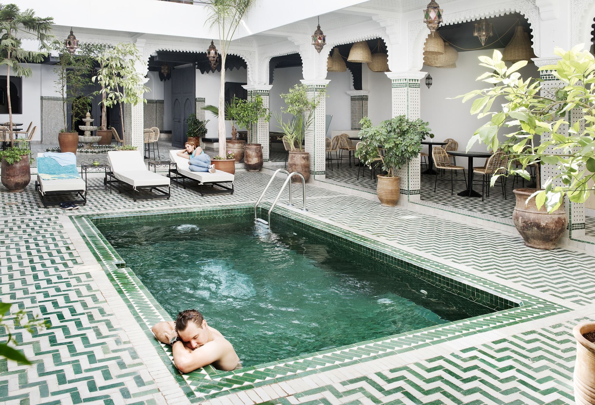 Una piscina con sillas de descanso y plantas en macetas, rodeada de muros y arcos blancos.