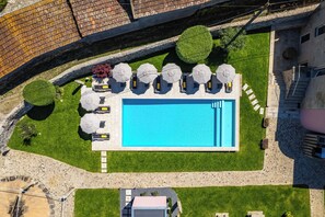 An aerial view of a swimming pool with lounge chairs and umbrellas, surrounded by well-manicured grass and stone pathways.