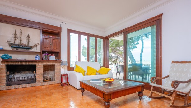 A spacious living room with a fireplace, a white sofa with yellow cushions, a wooden rocking chair, and large windows offering a view of trees. - Ca n Quiros in Mallorca (Santa Margalida)