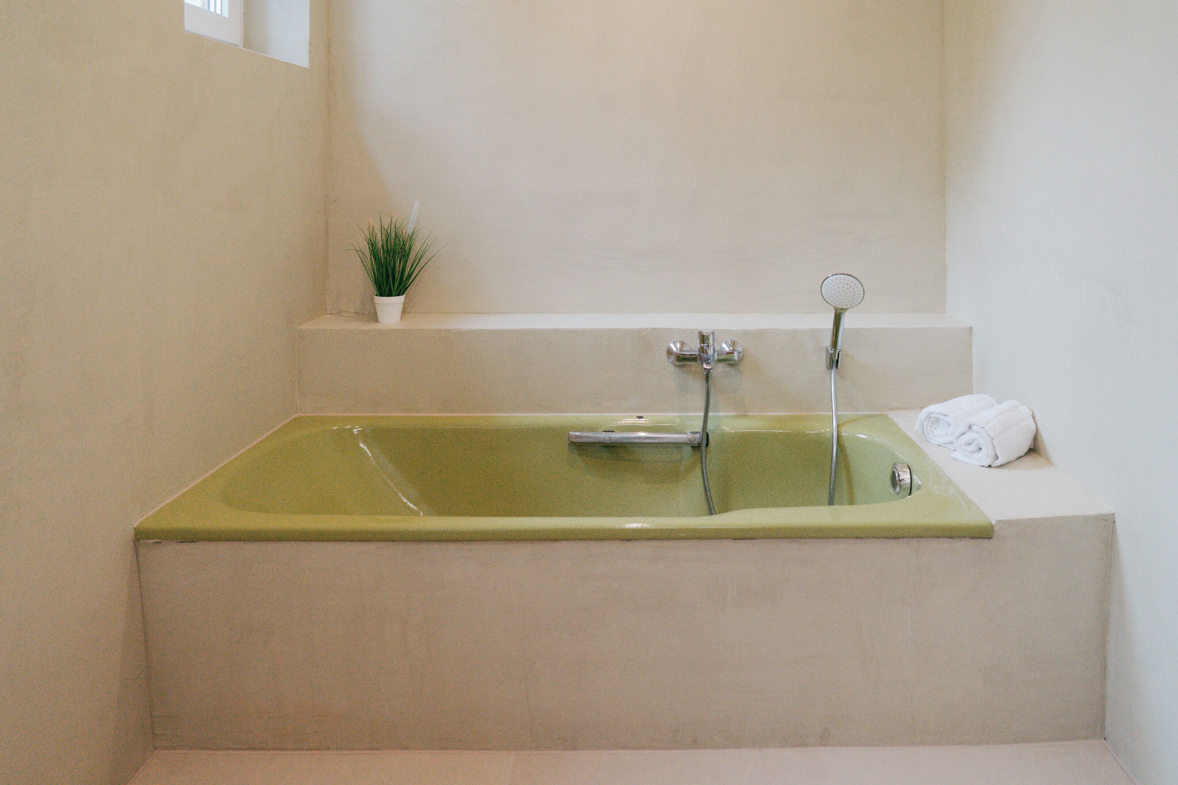 A rectangular bathtub with a greenish hue, a small potted plant on a ledge above it, and a rolled white towel on the adjacent wall.