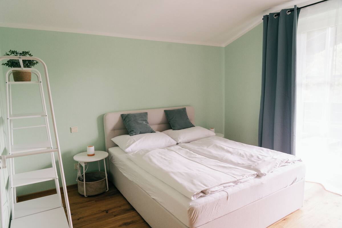 A neatly made bed in a room with a ladder, a small round table, and a potted plant.
