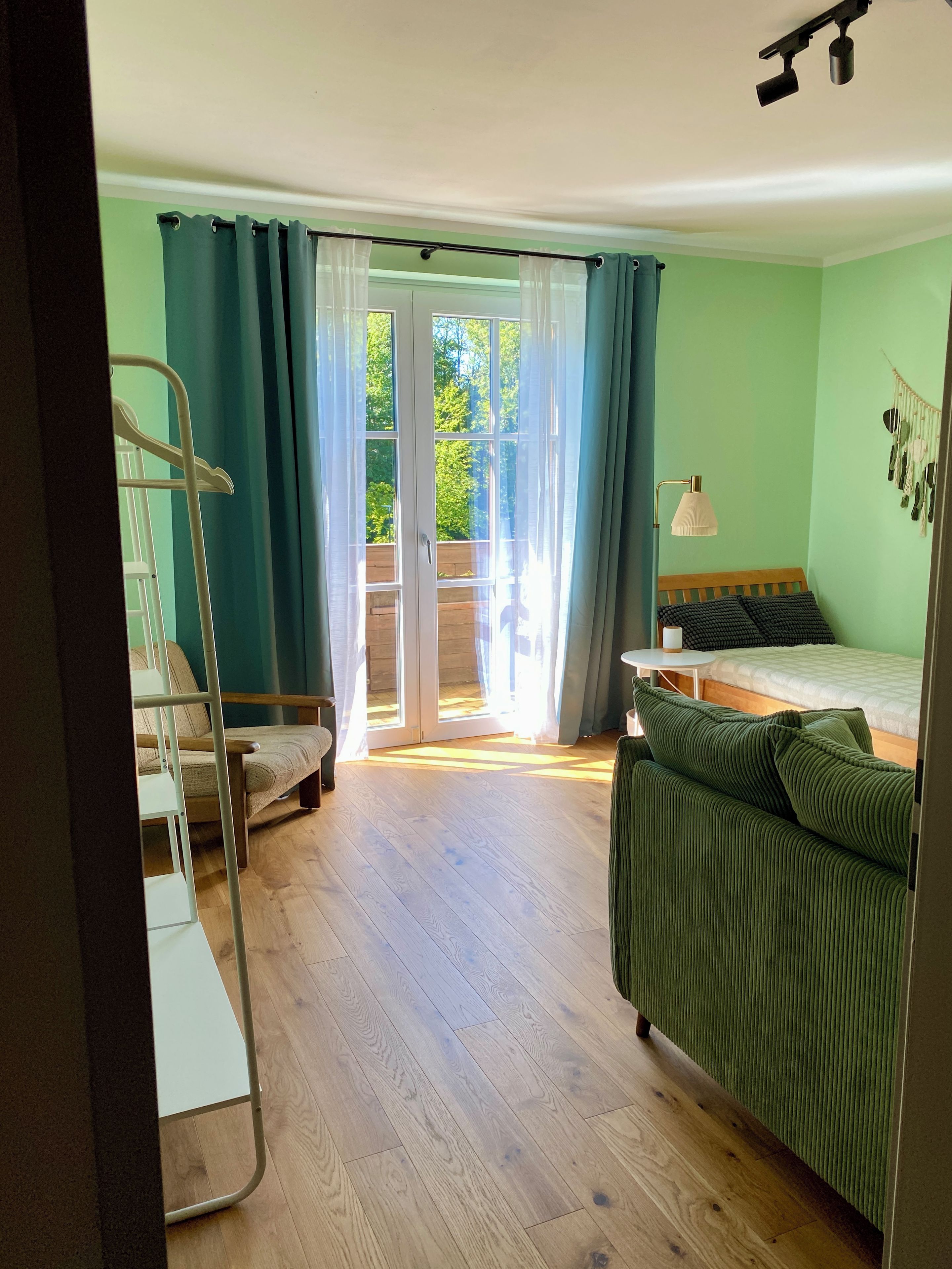 A room with a bed, a green sofa, a white lamp, a metal ladder, and a sliding glass door leading to an outdoor area.