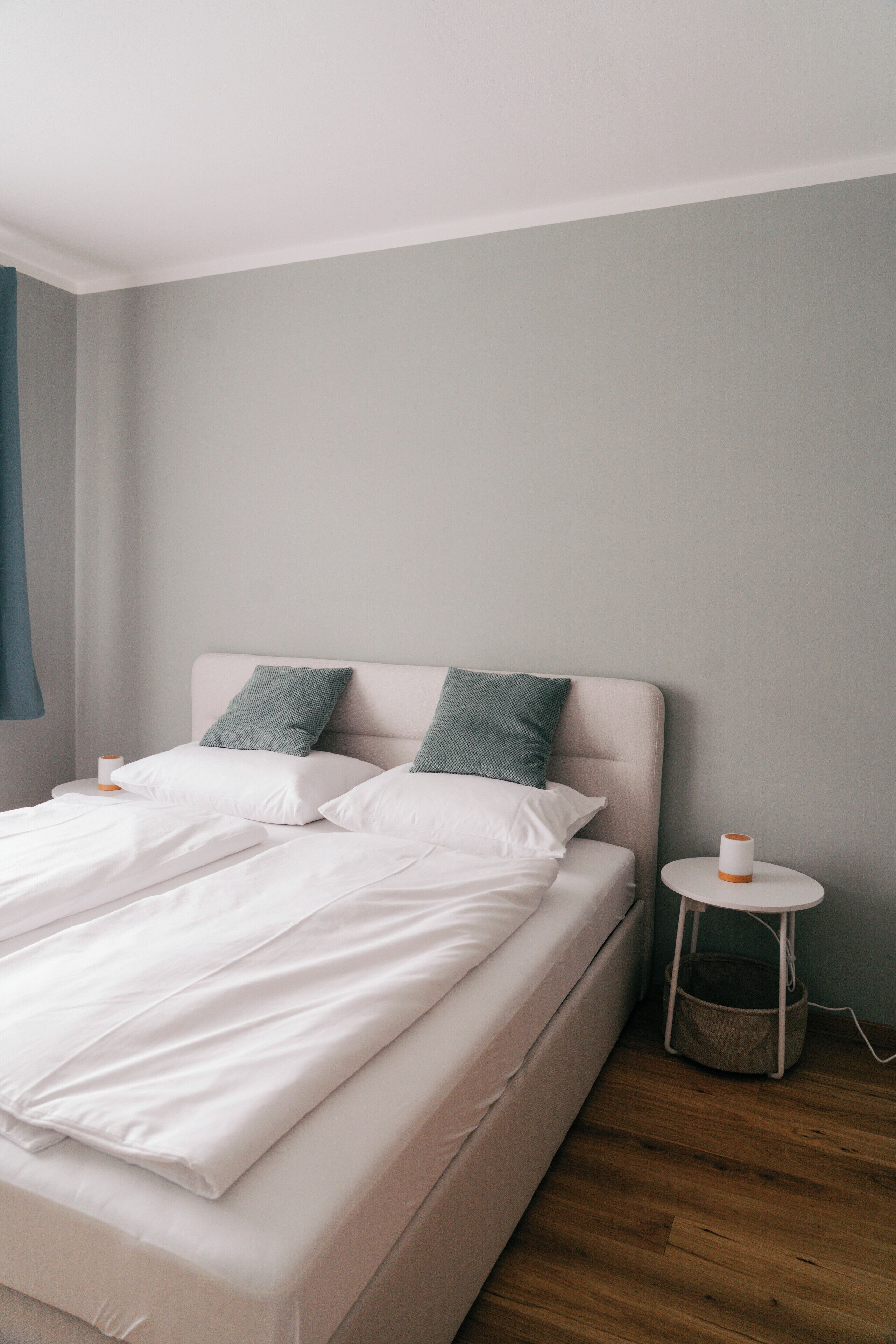 A neatly made bed with white linens and two green pillows, a small bedside table with a white lamp, and a wooden floor.