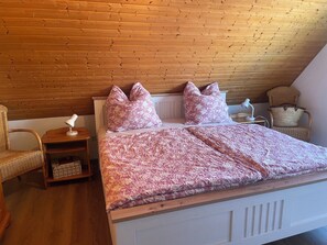 Une chambre avec un plafond en bois, un lit recouvert d’une couverture à motifs, deux oreillers, une table de chevet avec une lampe, une chaise en osier et une petite armoire en bois.