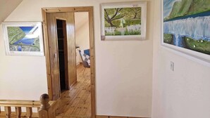 Ein Flur mit Holzboden, einer Holztür und einer Treppe mit Holzwand. An den Wänden hängen drei gerahmte Gemälde, die Landschaften zeigen.