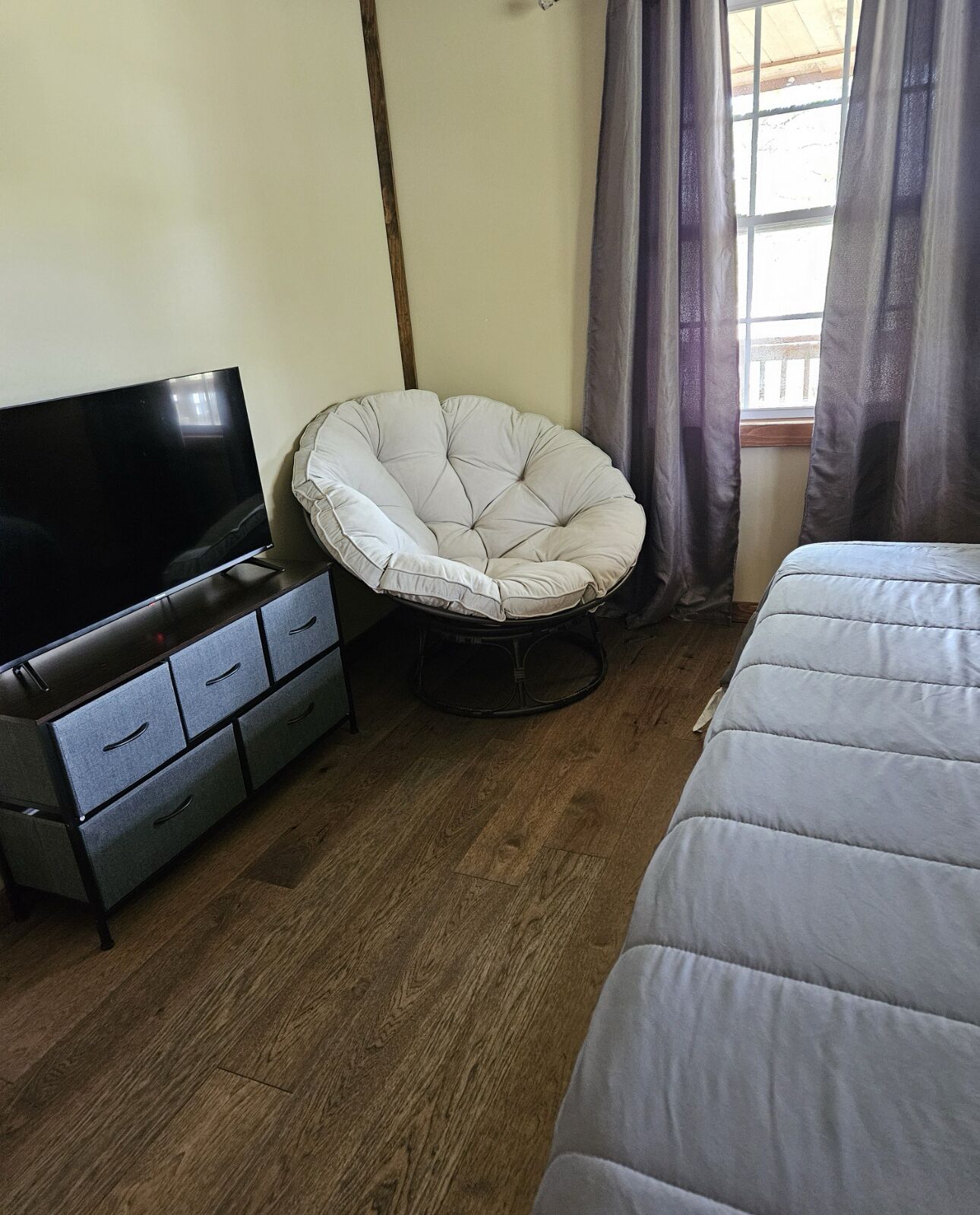 Une chambre à coucher avec un lit, un téléviseur posé sur une commode, une chaise et une fenêtre avec des rideaux.