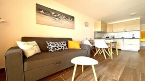 Una sala de estar moderna con un sofá gris, cojines estampados y un cuadro de pájaros. Hay un comedor con sillas blancas y una mesa de madera. La cocina tiene armarios de madera clara y electrodomésticos blancos.