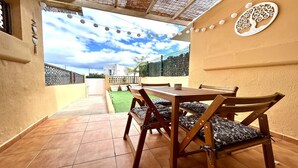 Un patio con mesa y sillas de madera, un mural decorativo con motivos vegetales y vistas a un césped verde y una cerca.