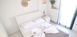 Una cama bien hecha con sábanas blancas, una mesita de noche con lámpara y una planta en maceta sobre un mueble blanco.