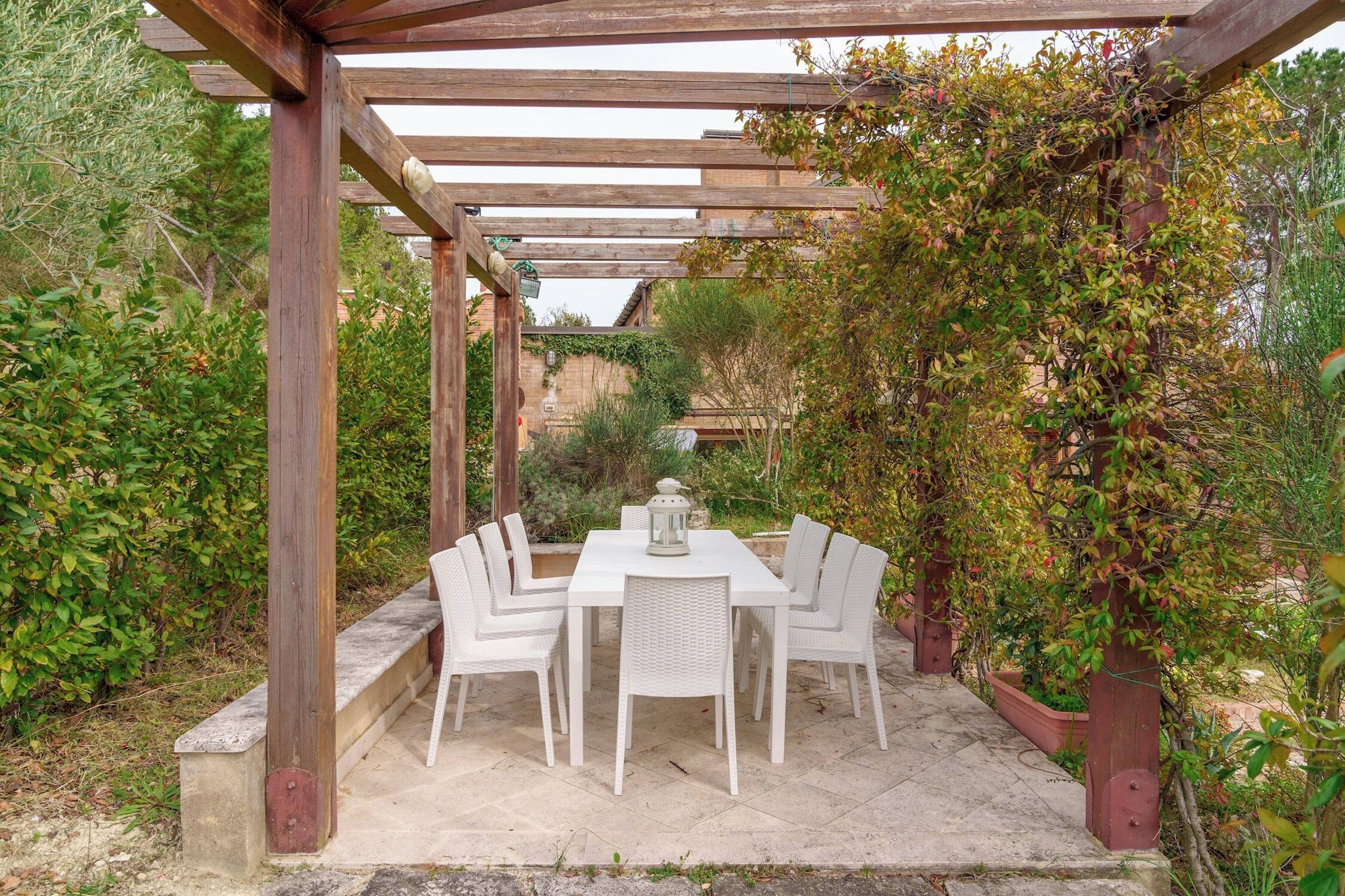 Et overdækket spiseområde med borde og stole, omgivet af grøn vegetation og en pergola.