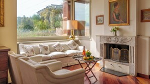 A living room with a sofa, armchairs, a fireplace, and a large window with a view of greenery. - Villa Lia 10, Emma Villas (Roccafluvione)