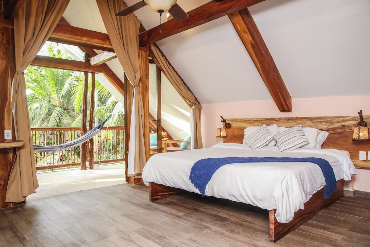 Un dormitorio espacioso con una cama grande, vigas de madera, una hamaca y vistas a exuberante follaje.