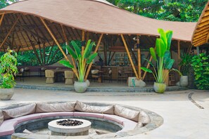 A covered outdoor seating area with a circular stone fire pit, surrounded by cushioned seating and large potted plants. - Masaya Tayrona & Beach Club (Santa Marta)