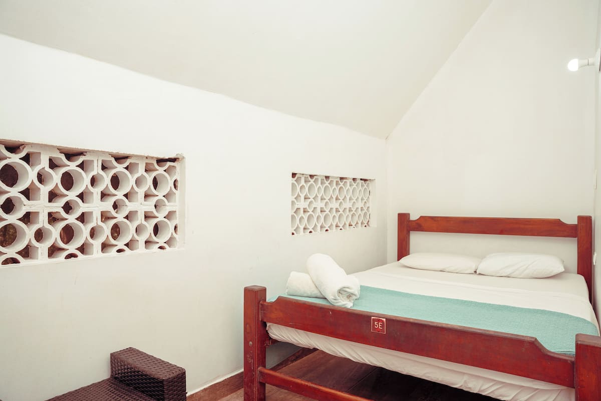 Una cama individual con estructura de madera, colchón blanco y manta verde azulada. Hay una toalla blanca sobre la cama. La habitación tiene paredes blancas y suelo de madera.