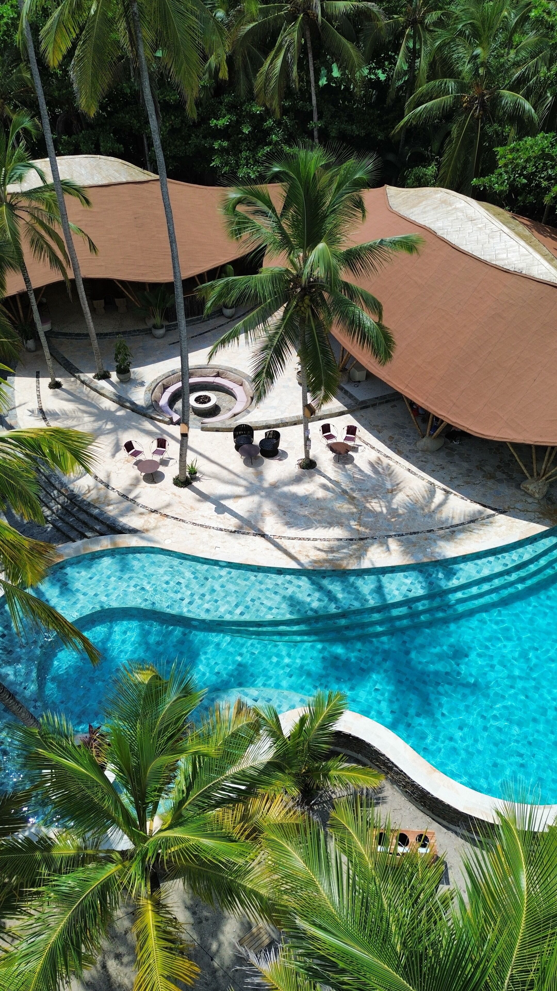 A resort pool area with a clear blue pool, surrounded by palm trees and covered seating areas.