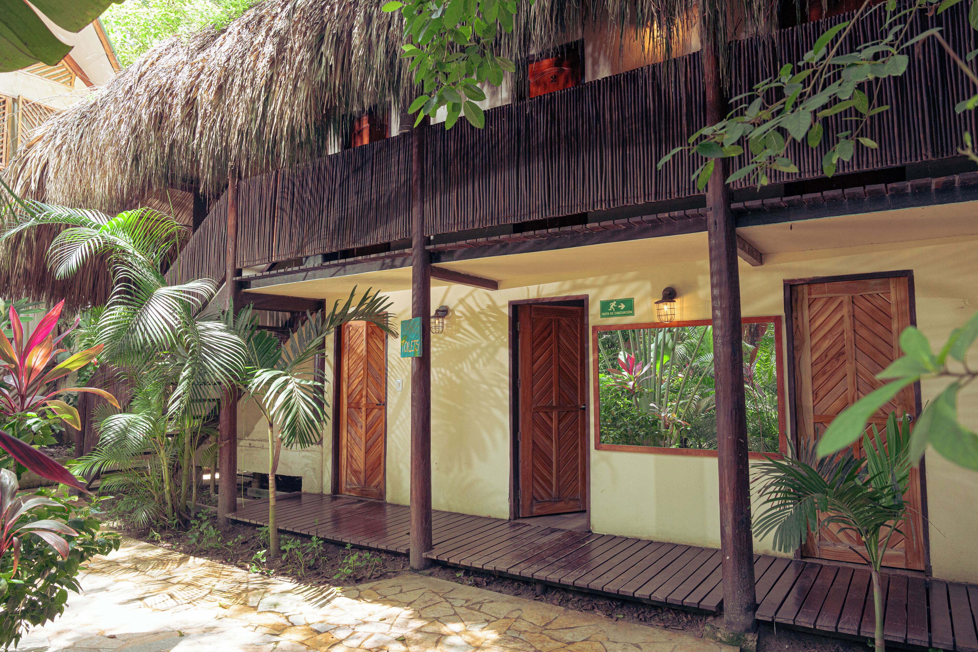 Un bâtiment à deux étages, doté de balcons en bois et de toits de chaume, entouré de plantes tropicales.