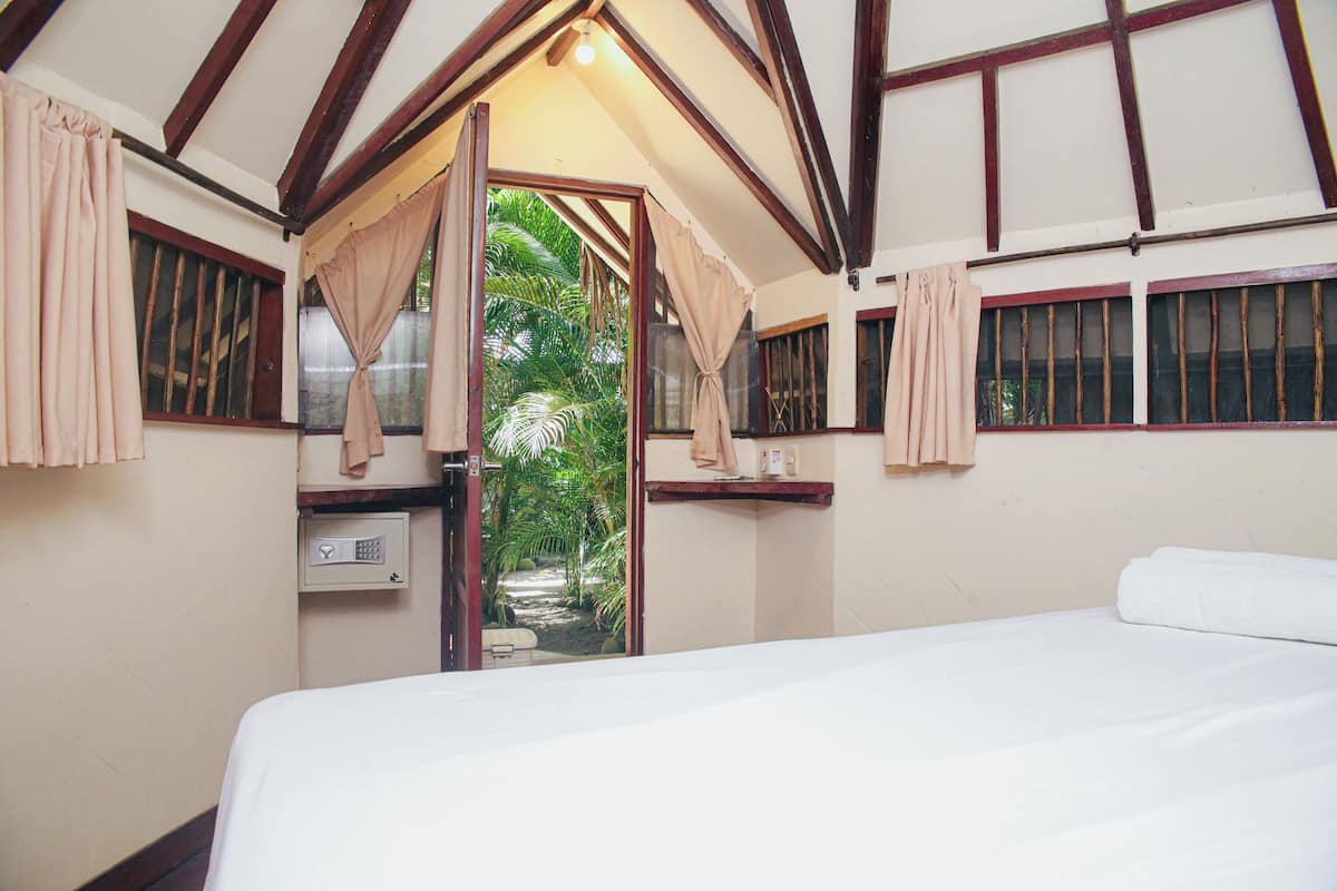 Un dormitorio con cama, ventana con cortinas, una mesita y vista a follaje exterior.