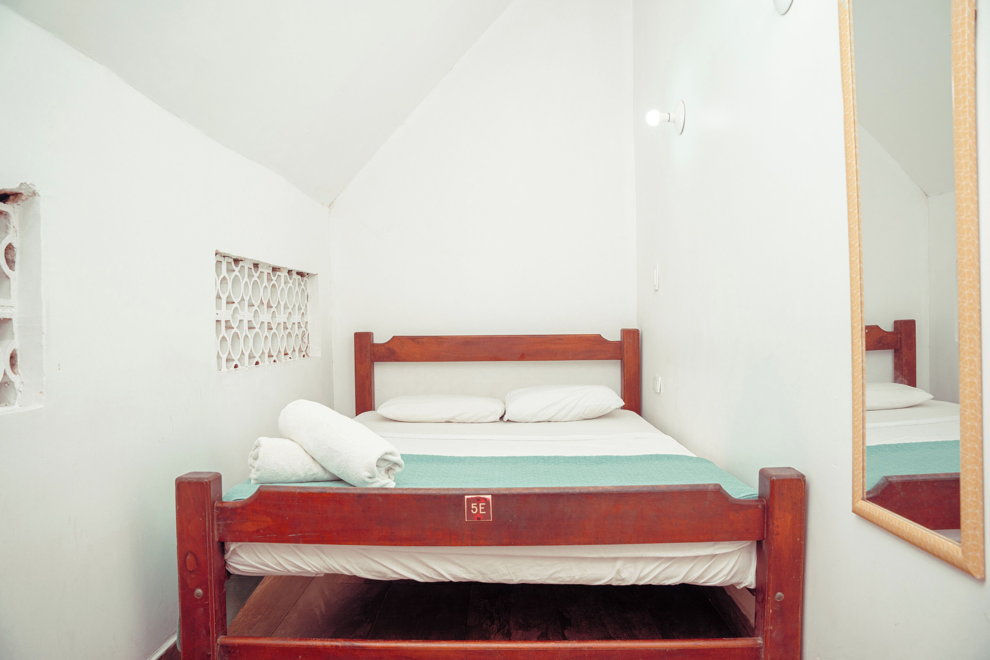 Una cama individual con estructura de madera, ropa de cama blanca y una ventana pequeña con un panel decorativo.