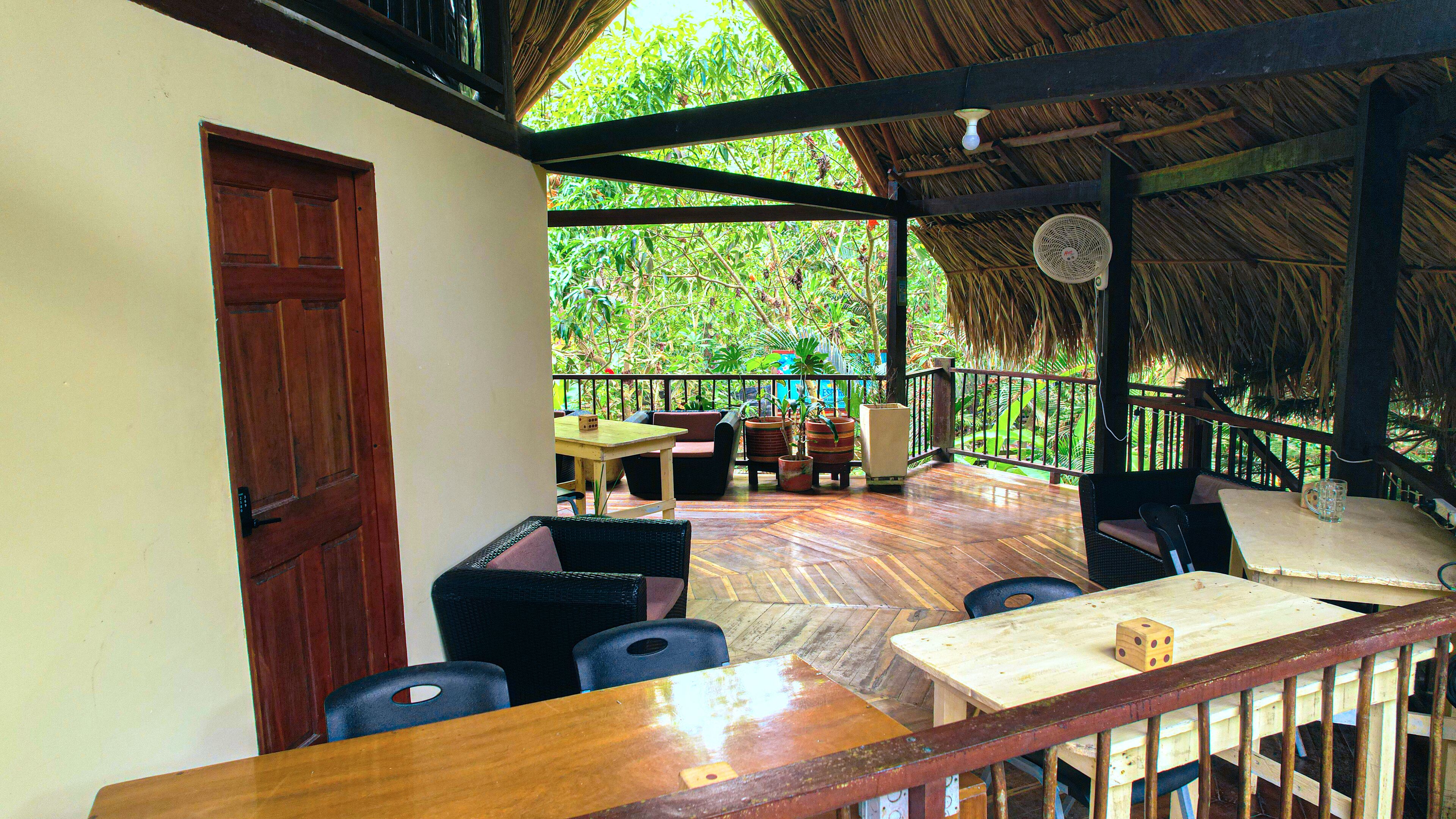 A covered outdoor seating area with wooden furniture, a thatched roof, and a view of lush greenery.