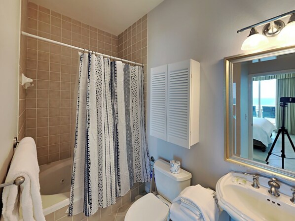 A bathroom with a shower curtain, toilet, sink, and a mirror reflecting a balcony with a camera on a tripod.