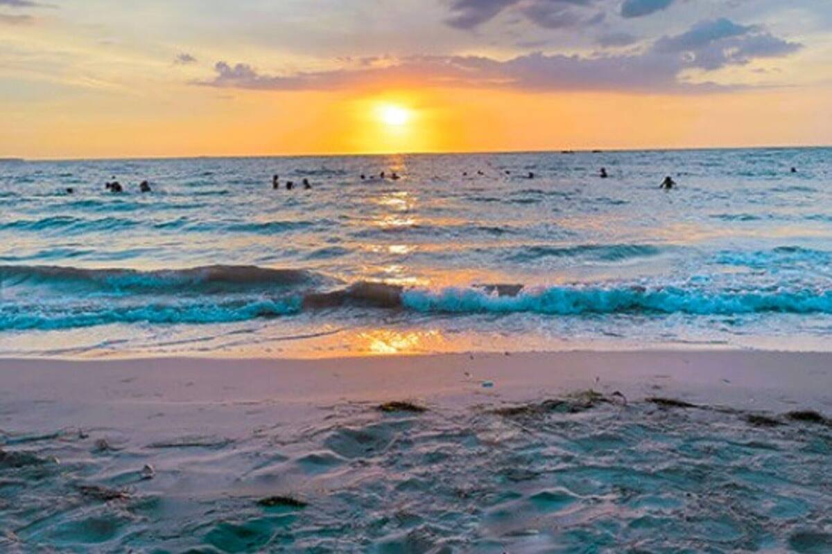 A beach with the sun setting over the ocean, waves gently lapping the shore, and people swimming in the water.
