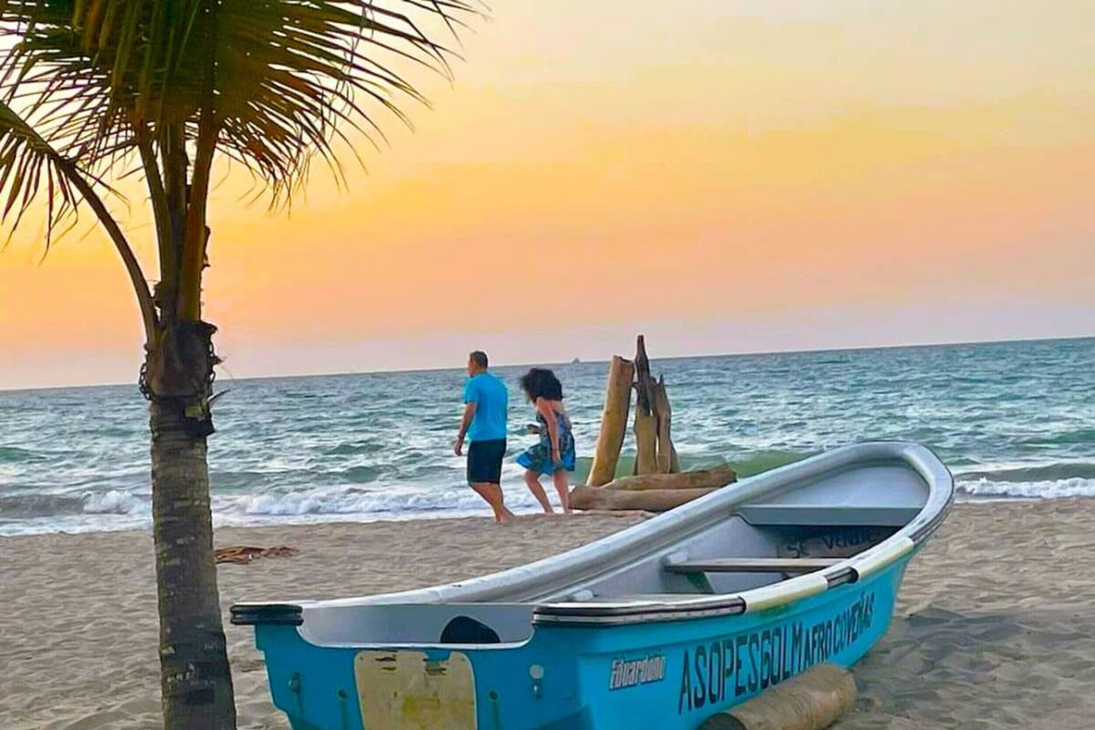 A beach scene with a blue boat named "ASOPESGOL MARINAS" on the sand, a palm tree, and two people near the water.