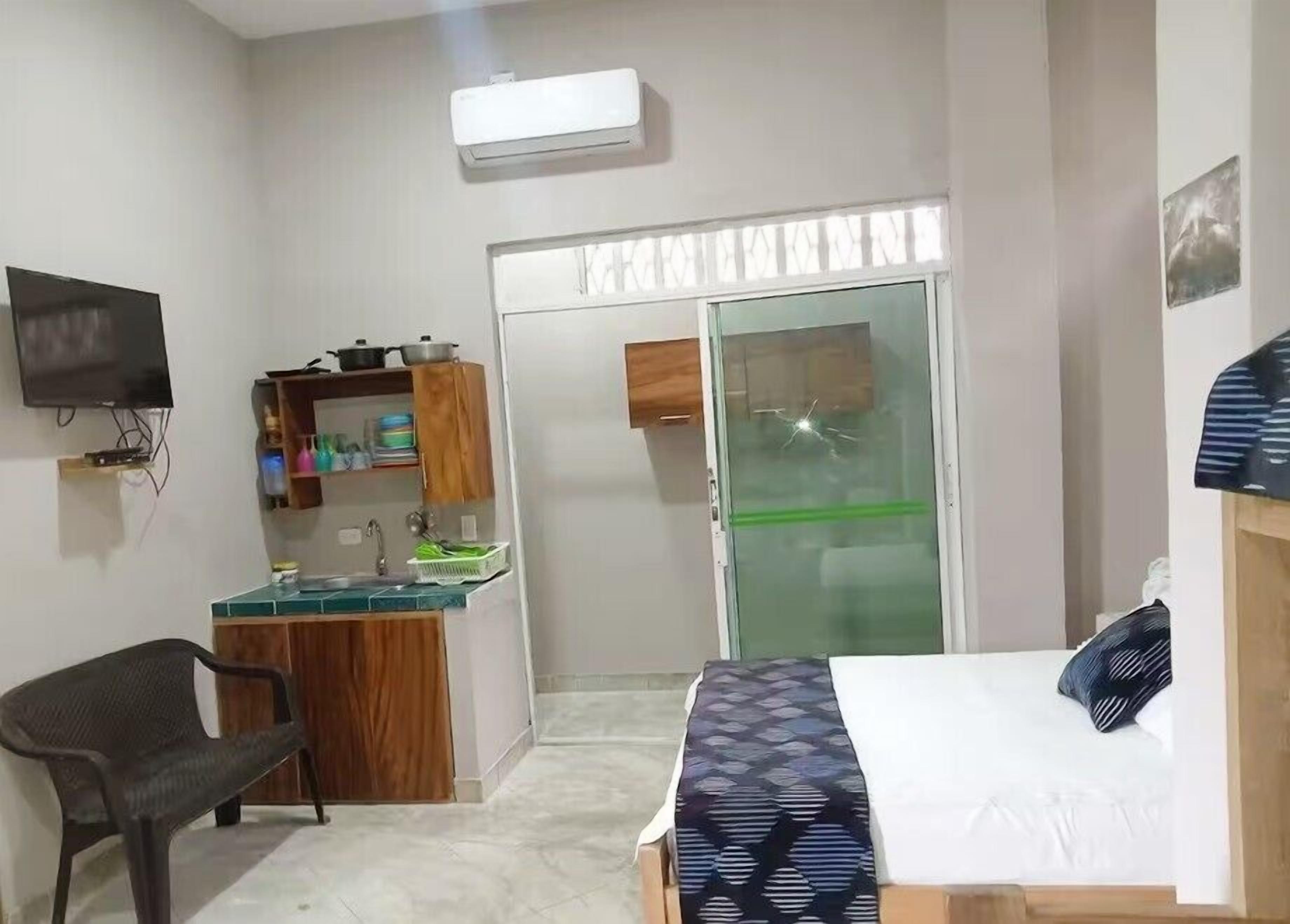 Un dormitorio compacto con cama, una silla, un pequeño rincón americano y una puerta corrediza de cristal que da a un baño.