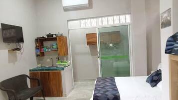 Un dormitorio compacto con cama, una silla, un pequeño rincón americano y una puerta corrediza de cristal que da a un baño.