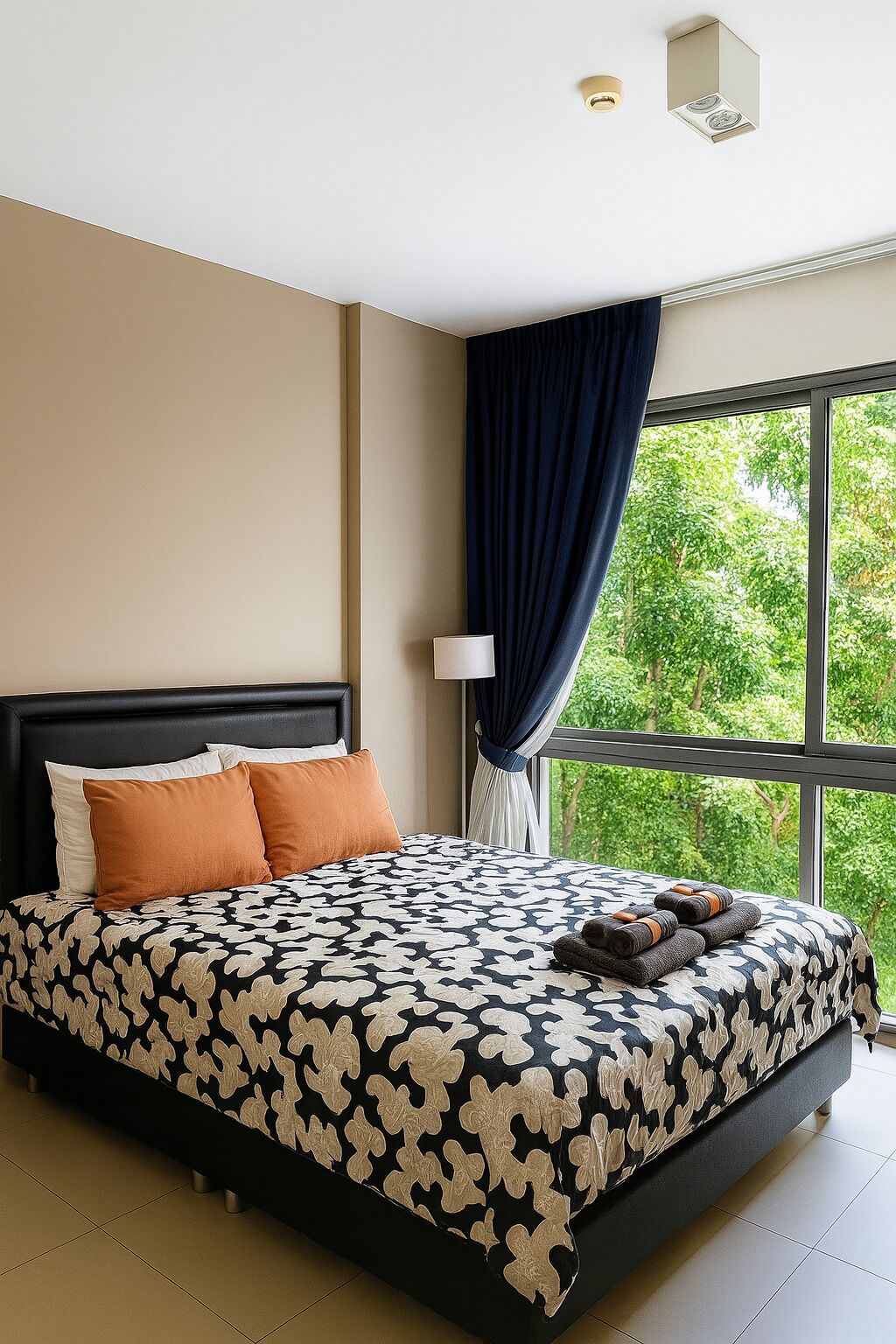 A modern hotel room with a large bed, a nightstand, a lamp, and a view of greenery through the window.