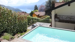 A swimming pool with a wooden deck, surrounded by greenery and overlooking a lake with mountains in the background.