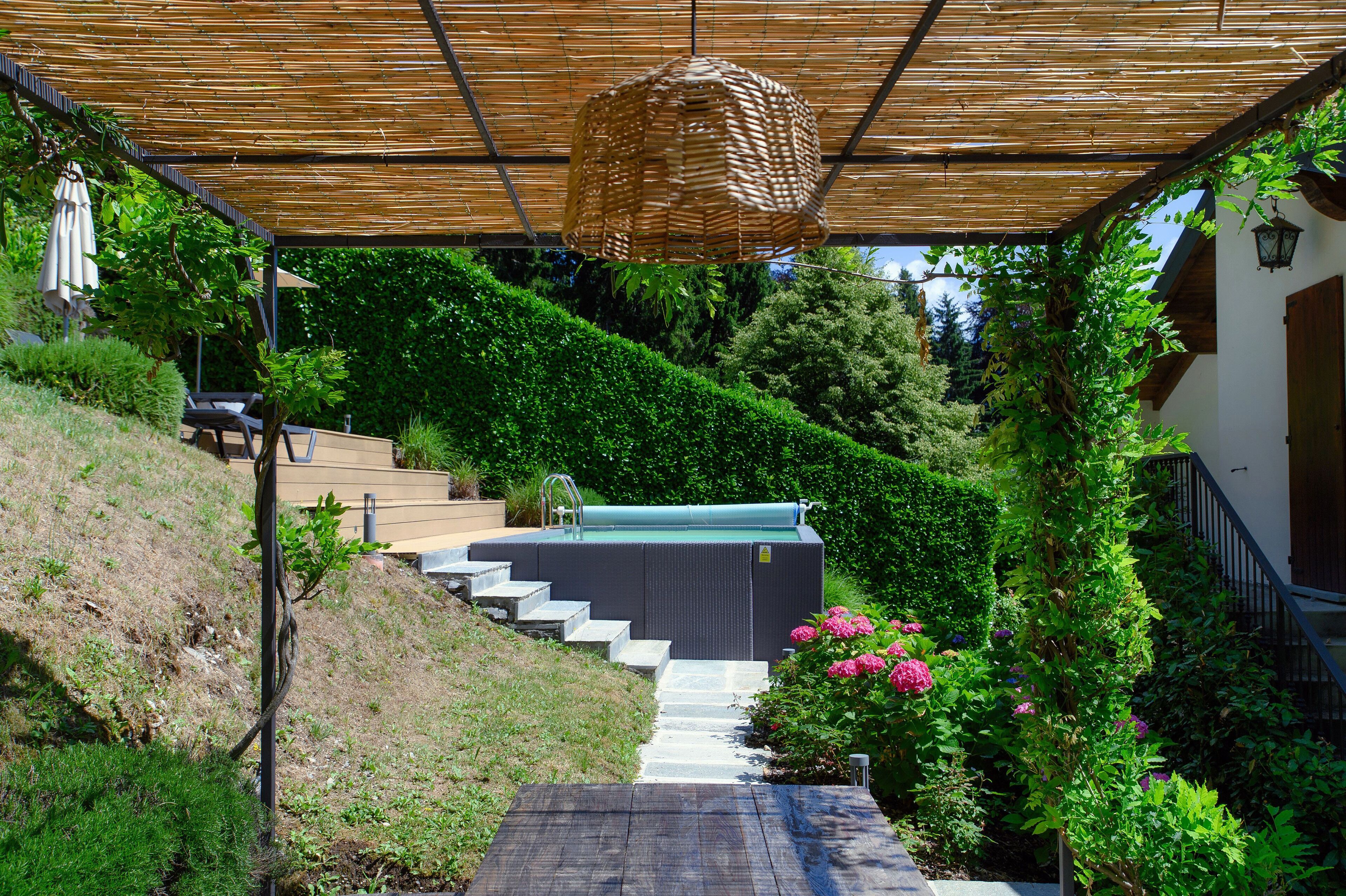 Et overdækket udendørs område med en trappe til en pool, omgivet af frodig vegetation og en træpergola.