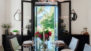 A dining area with a table set for four, a vase with flowers, and a view of greenery through an open door.