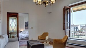 Ein Hotelzimmer mit Bett, Sofa, zwei Stühlen, einem Koffer und einem großen Fenster mit Blick auf die Stadt.