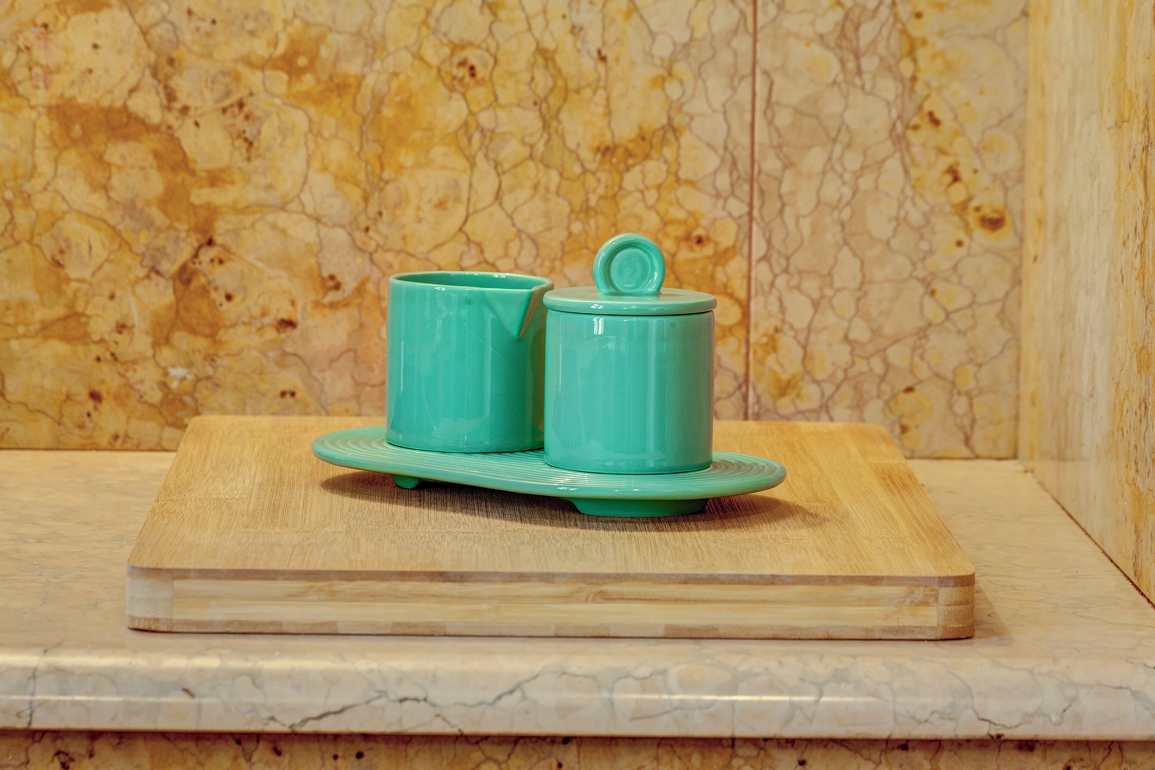 A wooden cutting board with a pair of teal ceramic cups and a lid on it.