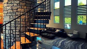 Ein modernes Interieur mit einer Steintreppe, einem Wohnbereich mit Sofa und Couchtisch sowie großen Fenstern mit Blick auf Grünflächen.