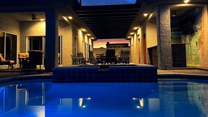 Ein Poolbereich mit Liegestühlen und einem Tisch, umgeben von Wänden mit strukturierter Oberfläche und Blick auf einen Sonnenuntergang.