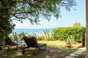Two wooden lounge chairs with cushions, a small table, and a potted plant in a garden setting with a view of the sea.