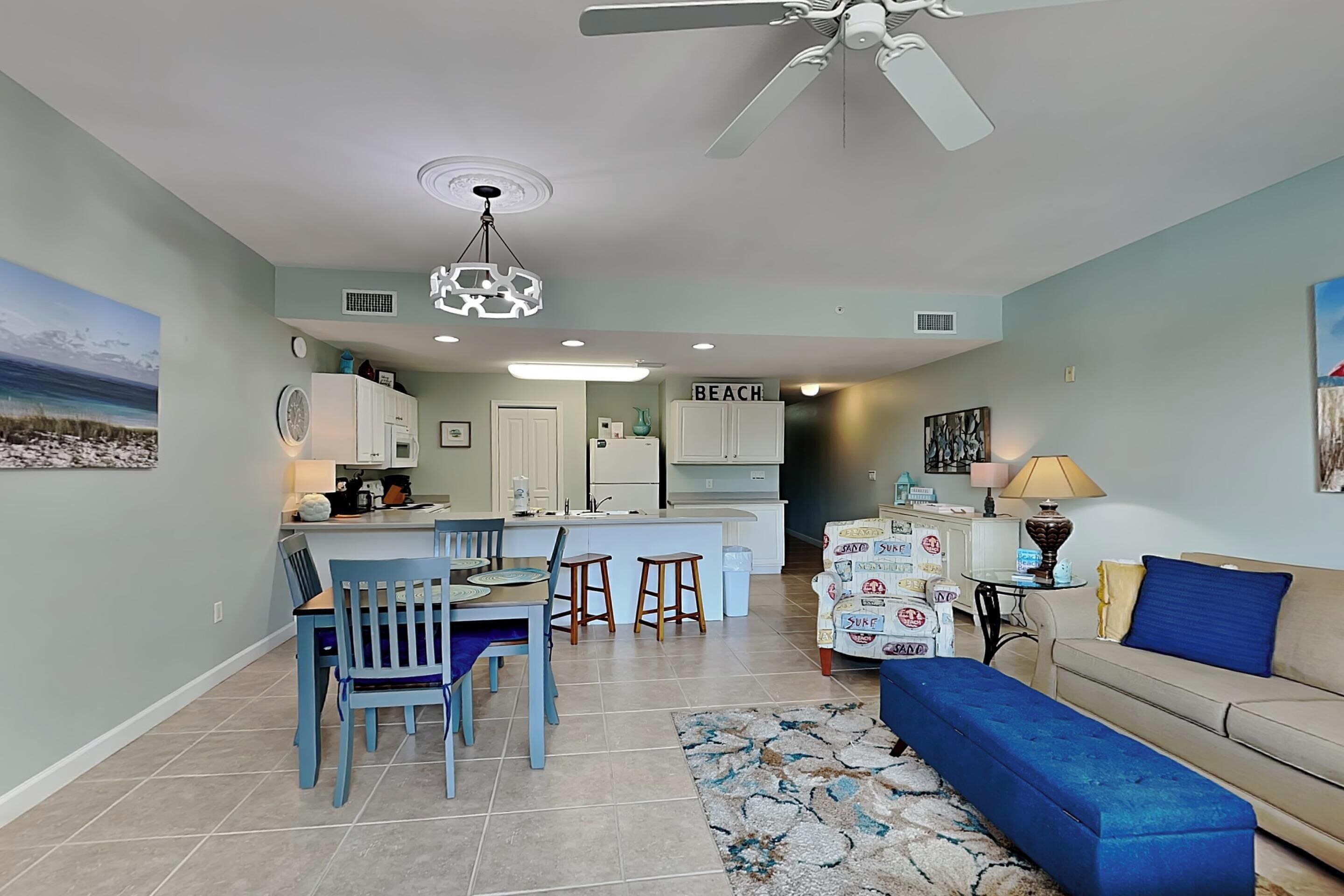 A modern kitchen with white cabinets, a dining area with blue chairs, a blue bench, and a beige sofa.