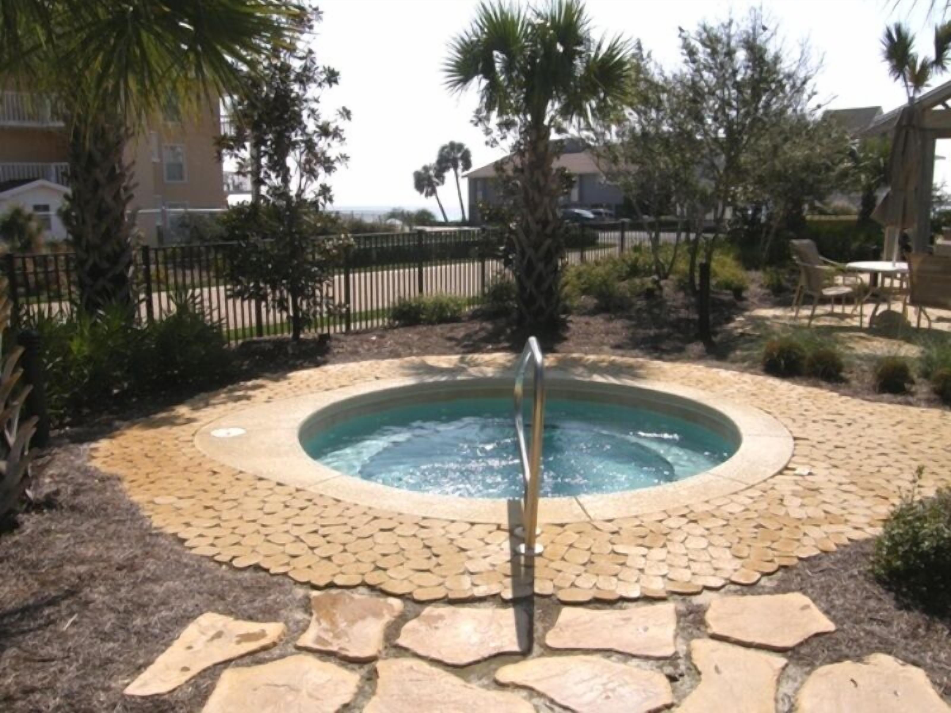 A hot tub surrounded by a stone border with a palm tree and a fence in the background.