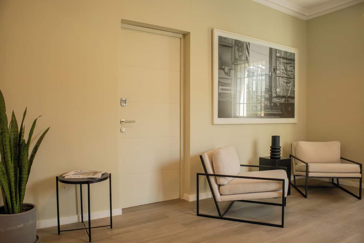 Una habitación con una puerta, una obra de arte enmarcada en la pared, dos sillas, una mesita con una planta, y otra planta en maceta.